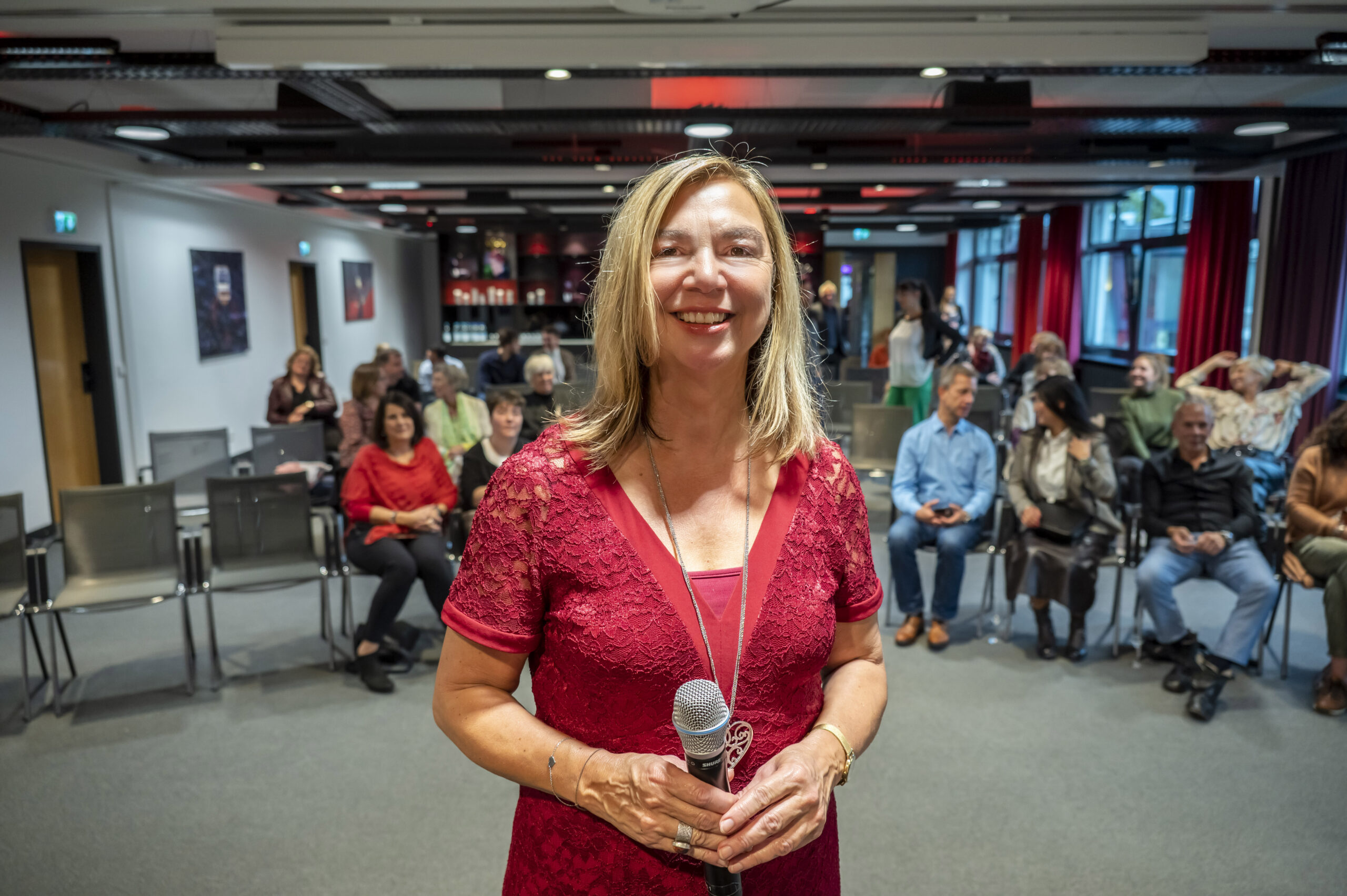 Eine Frau in einem roten Spitzenkleid mit einem Mikrofon steht lächelnd vor einem Raum mit sitzenden Teilnehmern. Das Publikum ist leger gekleidet, einige sitzen und andere schauen auf ihre Handys. Es scheint sich um eine zwanglose oder gemeinschaftliche Veranstaltung zu handeln.