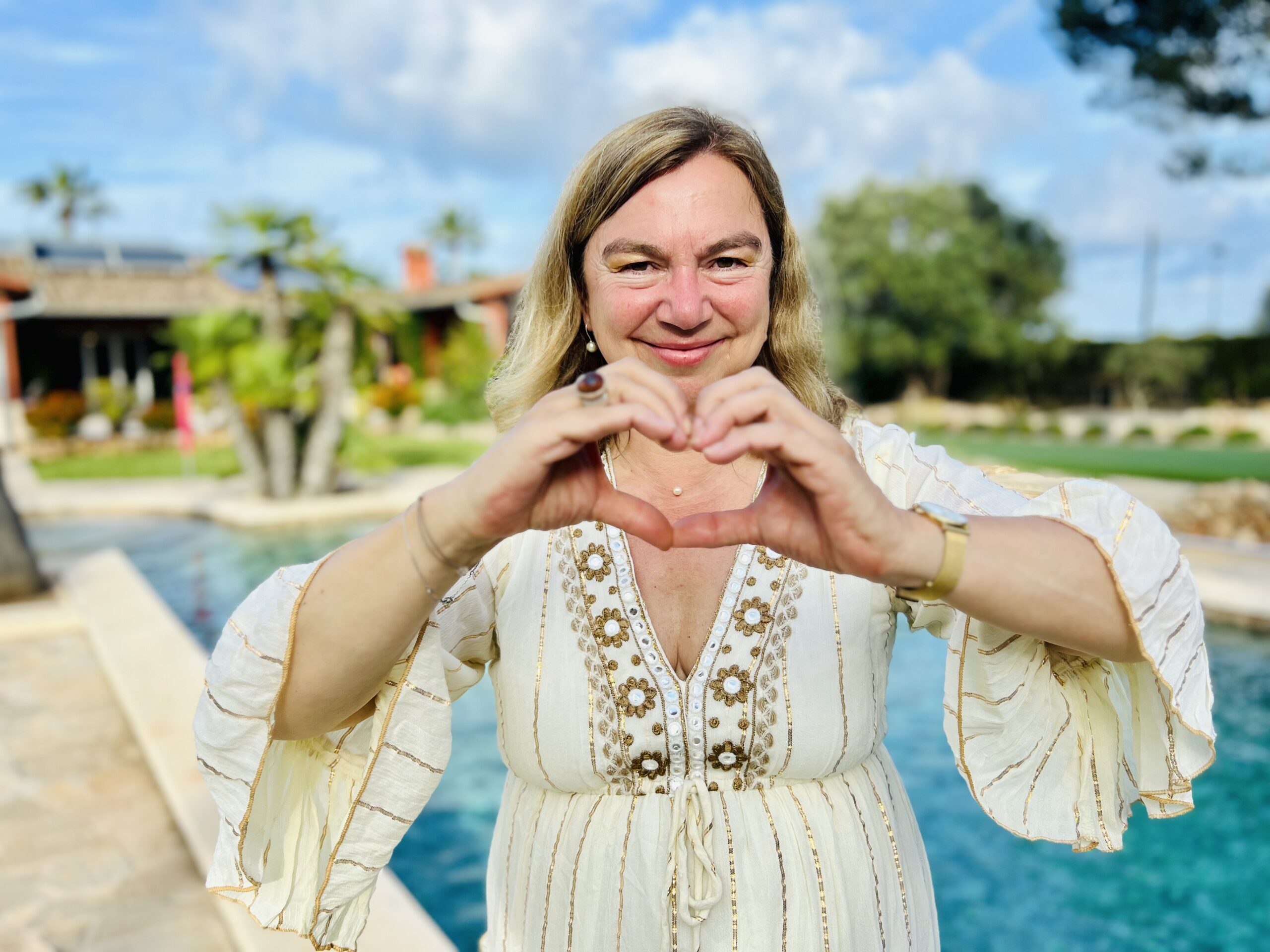 Eine Frau mit hellem Haar steht draußen an einem Pool und formt mit ihren Händen ein Herz. Sie trägt ein weißes, fließendes Kleid mit Blumenstickerei und lächelt in die Kamera. Im Hintergrund sind Bäume, ein klarer blauer Himmel und ein teilweise sichtbares Haus zu sehen.