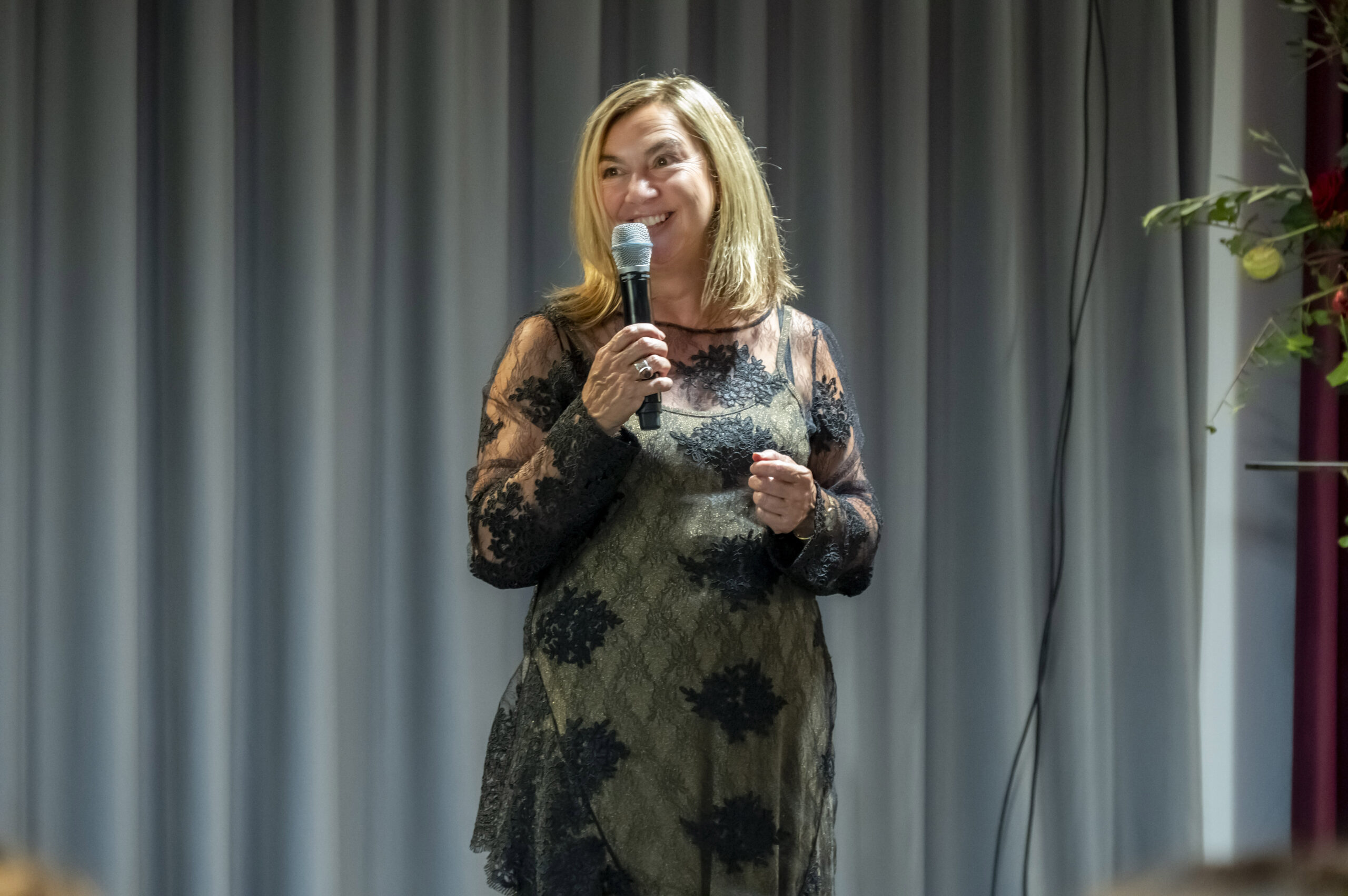 Eine Frau mit blondem Haar steht auf einer Bühne und hält ein Mikrofon. Sie trägt ein durchsichtiges schwarzes Kleid mit Spitzenmuster über einem dunklen Unterkleid. Im Hintergrund sind graue Vorhänge zu sehen und auf der rechten Seite ist ein Teil eines Blumenarrangements zu sehen.