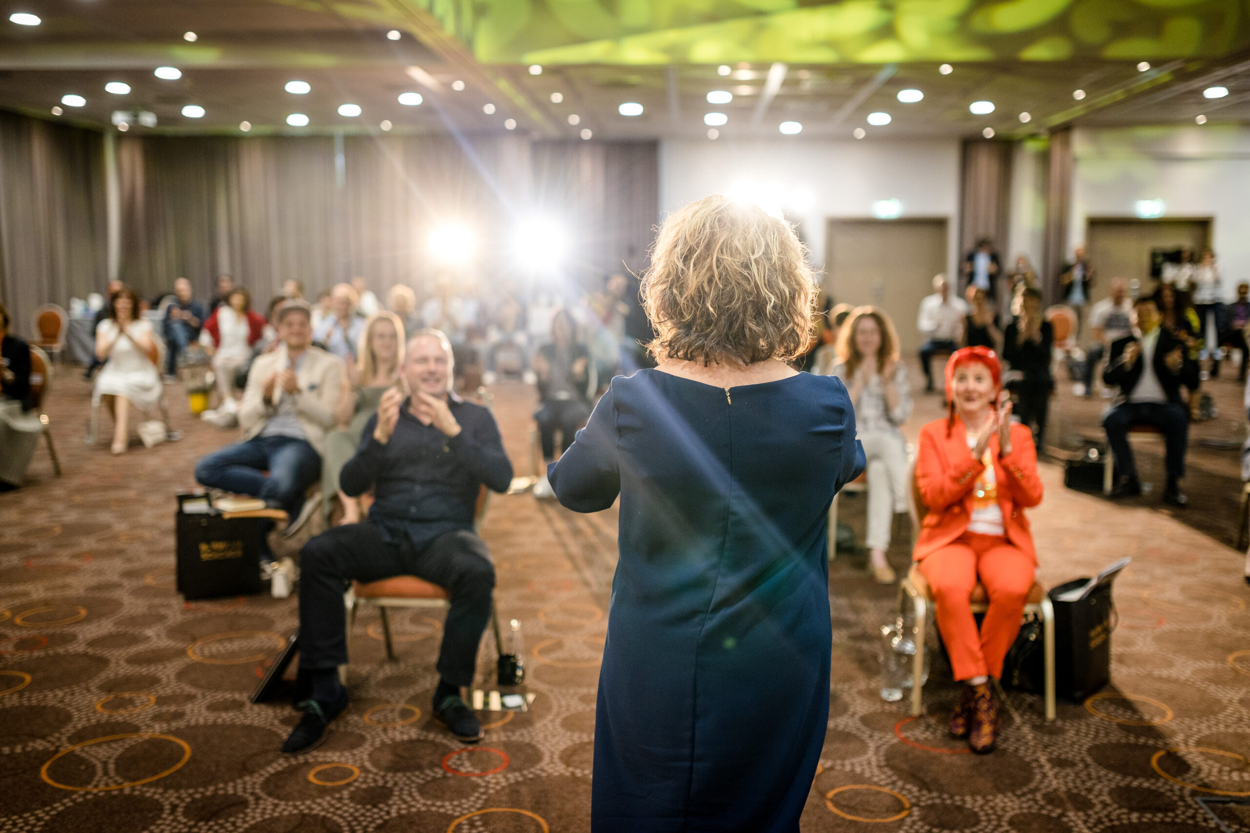 Eine Frau mit lockigem Haar steht vor einem Publikum und hält eine Rede oder Präsentation. Die Zuschauer, die auf Stühlen in einem großen, gut beleuchteten Raum sitzen, klatschen und wirken engagiert. Im Hintergrund sind einige Lichter zu sehen.