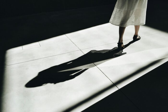 Eine Person in einem weißen Kleid und schwarzen High Heels steht auf einem sonnenbeschienenen Boden und wirft einen langen Schatten. Die Szene ist minimalistisch, mit starken Kontrasten zwischen den hellen und dunklen Bereichen auf dem Boden. Der Oberkörper der Person befindet sich außerhalb des Rahmens.