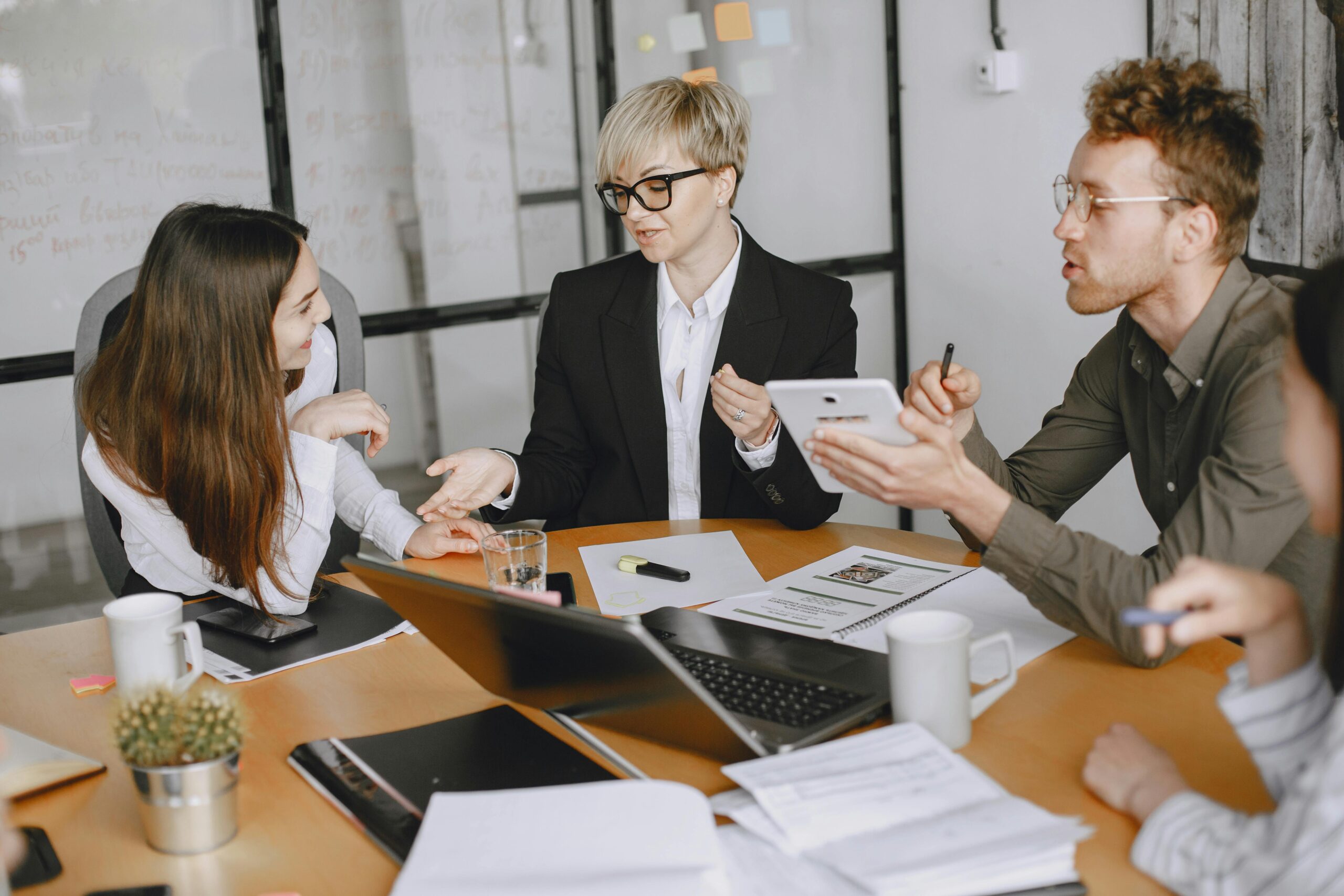 Eine Gruppe von vier Personen in einem modernen Büro diskutiert an einem Tisch mit Laptops, Papieren und Kaffeetassen. Eine Person hält ein Tablet, während eine andere mit der Hand gestikuliert. Im Hintergrund sind eine Topfpflanze und eine Glaswand mit Notizen zu sehen.