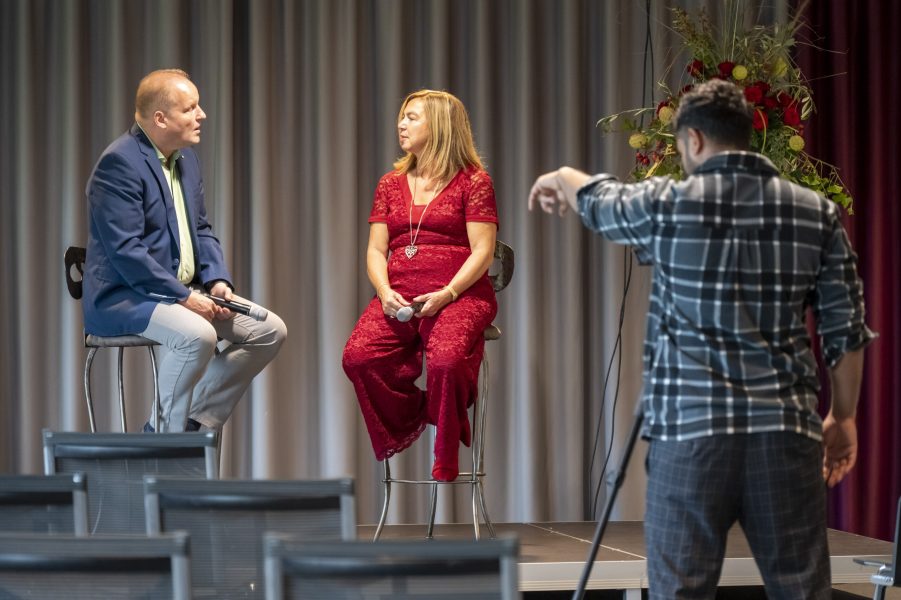 Zwei Personen sitzen auf Stühlen auf einer Bühne und unterhalten sich. Beide halten Mikrofone in der Hand. Ein Fotograf im karierten Hemd richtet seine Kamera auf einem Stativ aus und zielt auf sie. Hinter den Rednern ist ein Blumenarrangement zu sehen. Im Vordergrund stehen leere Stühle.